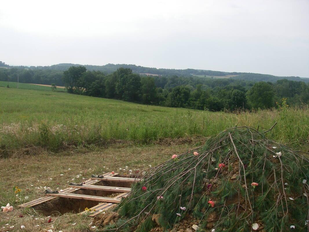 Open grave in a field of grass next to a pile of tree branches and flowers